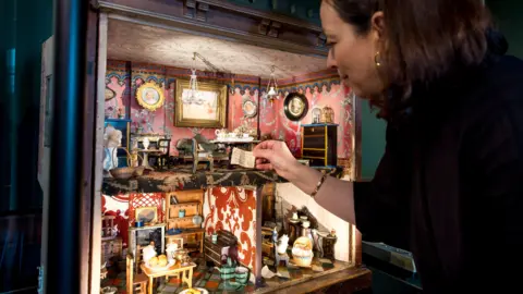 A woman with brown hair standing in front of the doll's house which is brightly coloured with three rooms across two storeys.