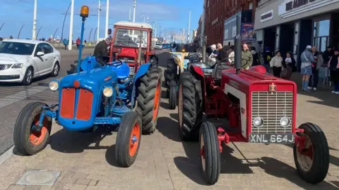A blue and red tractor stand side by side on the pavement. They have thin small old fashioned wheels on the front with big tredded wheels on the back. Two more can just be seen behind them.
