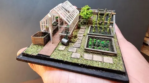 A miniature model of a garden with a greenhouse, vegetable patches and a gravel and paving slab path.