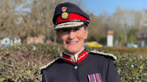 Anna Turner has tied-back fair hair under a formal uniform cap with a red band, and is dressed in black ceremonial uniform with white epaulettes, red collar detail and medals on the chest, standing outside and smiling into the camera.