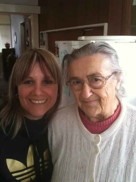 Andrea Urmston Two people positioned closely together in the foreground of a domestic kitchen. One wears a dark hooded sweatshirt with a gold logo; the other wears glasses, a white knitted cardigan with large buttons, and a pink top underneath. Behind them is a white refrigerator with magnets and notes attached. The background includes wood-panelled walls, a doorway leading to another room, and household furnishings. No bags, bins or floor markers are visible.