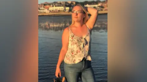 A photo of Karen Marks who poses by a waterfront. She has long blonde hair and wears a white floral tank top, jeans and she holds a pair of sandals in one hand while her other hand and arm is held behind her head. She is looking away from the camera as she smiles.