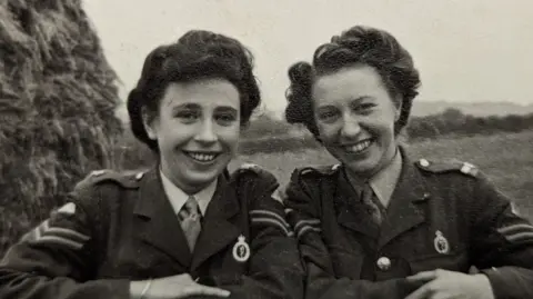 Joyce Goulborn Two smiling woman with dark hair, both wearing a shirt, tie and army jacket.