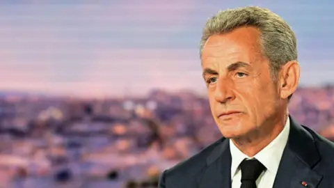 AFP via Getty Images Former President of France Nicolas Sarkozy looks off to the left of the frame. He is positioned in the far right. He is wearing a dark suit, white shirt and dark blue tie. His expression is slightly morose. The background is blurry and not quite clear. 