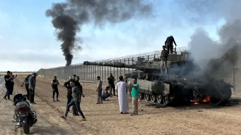 Reuters Palestinians climb on top of an Israeli military tank set damaged during Hamas's 7 October 2023 attack on Israel, on the Israeli side of the Israel-Gaza perimeter fence