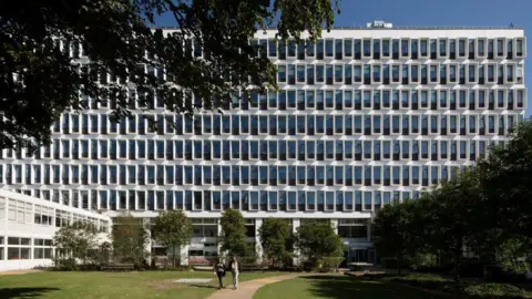 A large mid-rise building of white concrete and blue windows.The building facade is seen from a grass courtyard that also features multiple short trees.