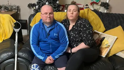 An image of two people sitting on a sofa, with Christmas decorations in the background. On the left is a bald man wearing glasses and a tracksuit jacket; his right leg is a prosthetic decorated with Saltire flags and a crutch leans against the couch next to him. On the right, a woman leaning against him, wearing a patterned shirt, with her hair in a bun.