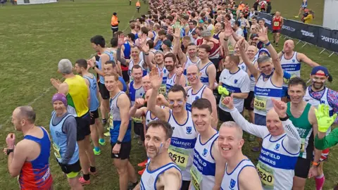 Chris Lines The start of the senior men's race with a large group of men in the white and blue Sedgefield Harriers vests looking into the camera and waving. They are standing in a grassy field and the camera is slightly above them.