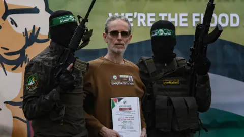 EPA Ohad Ben-Ami in a brown sweatshirt, standing between two Hamas fighters holding guns and with black balaclavas covering their faces. 