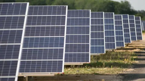 Nine solar panels lined up on a grassy field on a sunny day with hedges beyond. 