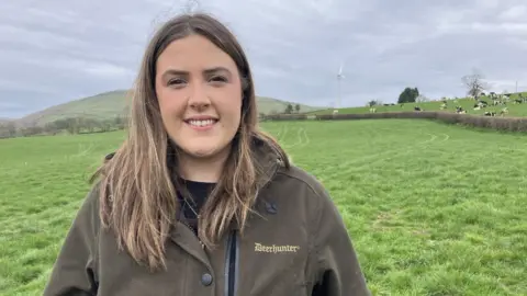 Elin Lewis stood in a field with cows in the distance. She has a brown jacket on and long blonde/brown hair and smiles at the camera.