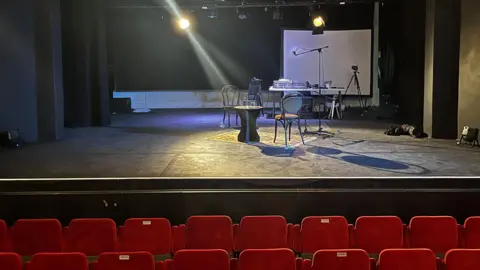 The inside of a theatre, showing the stage. There are red seats at the front. On the stage is a table and chairs. Several lights shine into shot.