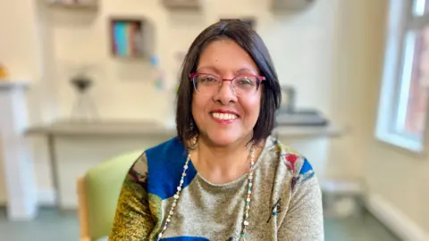 Andrea Thasan looks directly at the camera. She is wearing red-metal trimmed glasses and is wearing red lipstick. She has black hair cut into a bob swept to the side. Thasan is wearing a long-sleeved multi-coloured top and has a string of beads on. She is smiling. Thasan is sitting on a green chair in a training room with a fireplace and a cream wall. The background is slightly blurred.