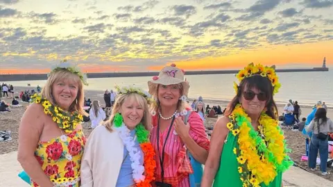 The women are wearing flowers in their hair, on their hats and around their necks as they stand in front of the sea at sunrise. The sky is coral with small grey clouds and there is a lighthouse on a harbour wall across the water behind them.