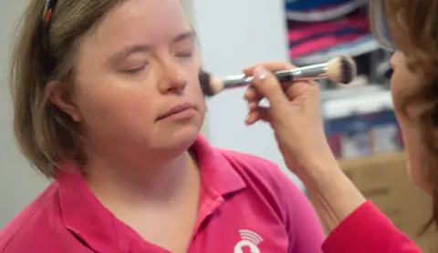 Emily Ann Riedel sminker ansiktet før hun går foran kameraet. Hun lukker øynene mens en makeupartist sminker seg med en børste. Hun har ikke på seg brillene og har på seg en rosa poloskjorte.