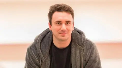 Sam Smith Sam Smith is looking at the camera. He is wearing a grey fleece hoodie and black tee shirt and has short brown hair.