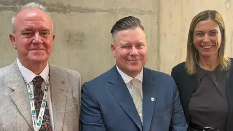 (Left to right) A man with a grey suit, white shirt and tie with a North Northamptonshire Council-branded white and blue lanyard round his neck. A man in the middle with a blue jacket, white shirt and tie wearing a pin badge. A woman with a black jacket and brown dress with belt. All three are looking directly at the camera.
