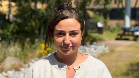 A photo of Meena in a grey cardigan with her brown hair tied up, standing outside the Queens Medical Centre in Nottingham.