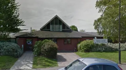 A street view image of the practice, a red-brick single-storey building surrounded by shrubs and trees. A sign that reads "Dinnington Group Practice Medical Centre, late night pharmacy" sits near the building entrance.