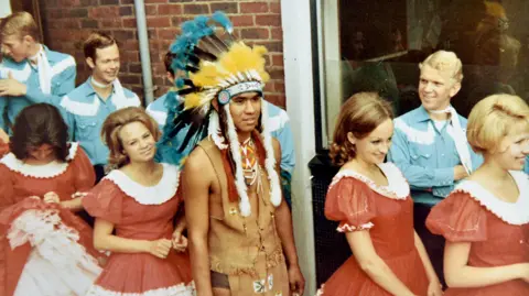 A 1960s picture showing several young women in red and white dresses with young men in background wearing light blue shirts and talking to each other. They are lined up alongside a brick building. A young man in middle of photo is wearing a Native American outfit with traditional headdress. 