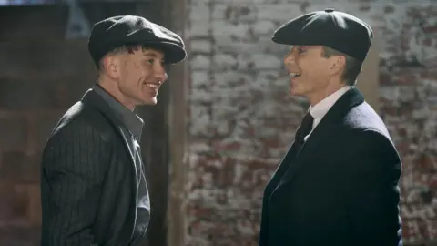 PA Barry Keoghan and Cillian Murphy, in the upcoming Peaky Blinders film. They are laughing and both are wearing dark suits and caps.