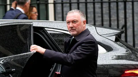 PA Media Steve Reed getting out of a rain-speckled black 4X4 vehicle in what looks like Downing Street. He is wearing a dark blue raincoat, and has neatly trimmed grey stubble and short salt and pepper hair. His left hand is holding the rear left door of the car, which is open, as if Reed has just got out and is about to shut the door. He is looking off camera to the left of frame. Behind him are black metal railings that are out of focus. There are two people apparently standing talking to each other next to the railings, but behind the vehicle.