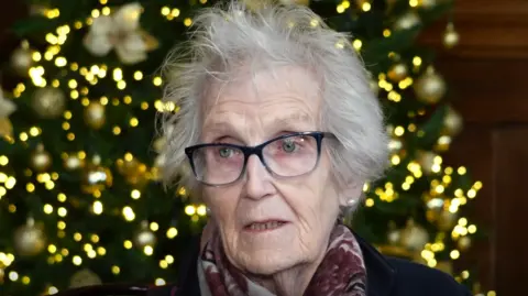 Ivy Wells is in the middle of the image. She has short grey hair and is wearing glasses and a black coat. She is sat in front of a Christmas tree. 