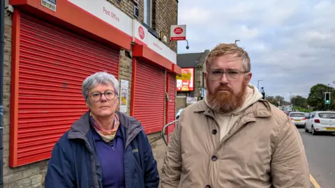 Submitted Two people standing in front of a post office branch with the shutters down. They both look very serious. The branch is on a busy road with lots of traffic passing. The man is middle aged and has ginger hair and a beard. He wears a beige jacket. The woman is older and had cropped greying hair. She wears a blue zip up jacket. 