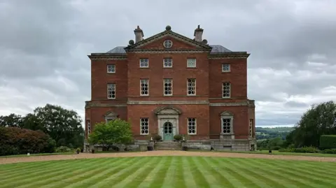 A red-brick stately home with more than a dozen windows at the front stands ahead of a green, mown lawn. Overhead are cloudy skies