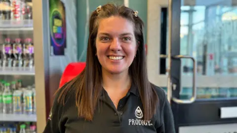 Sophie Corlett has long straight brown hair, clipped at the top to pull it away from her face. She is smiling and wearing a black polo shirt that says "prodigy" on it. She is standing by the door of a cafe on the right, with a drinks fridge on the left.