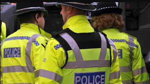 BBC Two male and one female police officers stand with their backs to the camera. They all have florescent jackets on and black helmets.