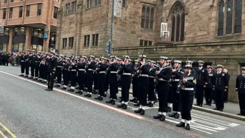 Crewmen from HMS Prince of Wales stand to attention on the road outside the church, wearing full uniform with white caps and holding their rifles