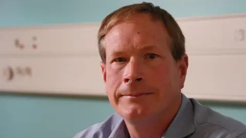 Dr Mark Weatherhead has short, dark blonde hair and is clean shaven. He is sitting in a consulting room at Wansbeck Hospital in Northumberland with a blue wall and white panel for instruments behind him. He is wearing a lavender blue shirt.