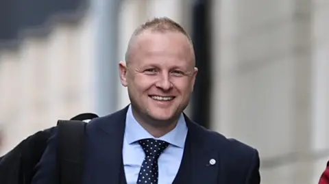 PA Jamie is smiling at the camera. He has blond spiky short hair and blue eyes. He is wearing a navy suit jacket, blue shirt and navy tie with white spots on it.