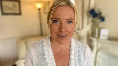 A woman in a white top smiles at the camera. Behind her, out of focus, are a chair, lamp and flowers.