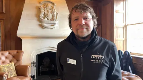 Man standing in a wood-panelled room beside a stone fireplace with a carved crest, wearing a black jacket labelled “Boleskine House” and a name badge, with leather chairs and a window letting in daylight behind him.