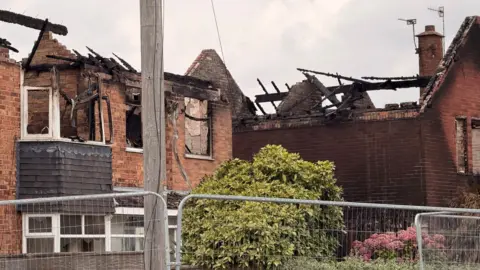 Two houses, with collapsed roofs, can be seen behind metal fencing. There are gardens in the foreground and a telegraph pole.