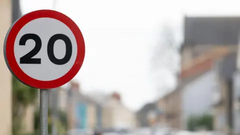 A circular 20mph sign. A blurred residential street can be seen in the background.