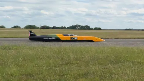 A photograph of the JCB vehicle on a runway. It is a long vehicle with a yellow front with "JCB" on it. The back half of the car is dark green and black with white writing. To the front and back is grass and it is on a grey surface. 