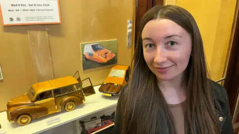 John Devine/BBC Kateryna Vidiushenko, 26 has long dark hair parted high on her forehead she is wearing a black leather jacket over a light brown shirt and behind her are some of Mr Clarke's wooden and brass models displayed in a case in the museum.