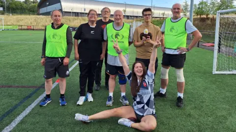 A group of men and women in yellow bibs and sports clothes pose together on a 4G sports pitch.