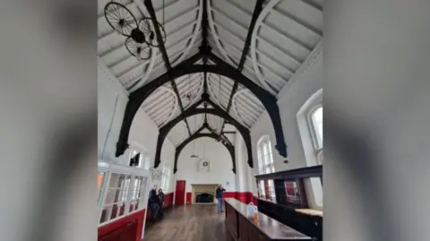 The empty waiting room is long and narrow. It has a bar on the right of the photo and large, almost floor-to-ceiling windows on either side. The walls are painted white, with some red features. The high vaulted ceiling is painted white with some arches painted in a dark colour as a feature. There is a large fireplace at the end of the room and three people are standing in the empty space.