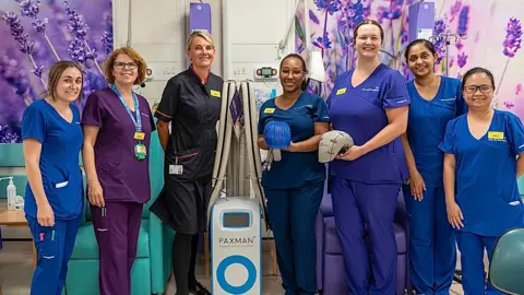 Surrey and Sussex NHS Trust A group of NHS staff in clinical uniforms standing either side of a newly donated scalp cooling machine