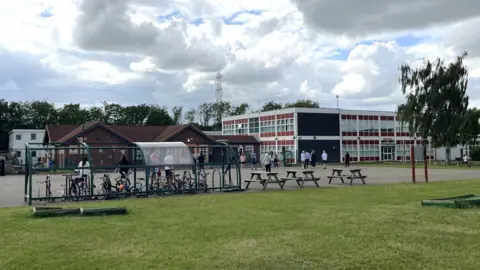 Hazlewood Primary School. There is a span of grass, then a concrete playground with a bike rack and picnic tables in front of the school buildings themselves. Two large buildings are visible. One, which appears to be older, is a single-storey building with a slanted roof. The other is a modern two-storey building with rows of glass windows on every side and a flat roof. Parents wait outside for their children to finish school.