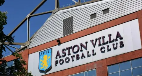 Getty Images On a sunny day with a tree to the left of the shot, a large sign has an AVFC badge and Aston Villa Football Club next to it. It is attached to the outside wall of the Aston stadium.
