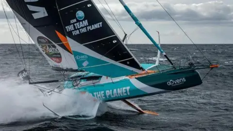 Pierre Bouras A racing yacht breaching the water with foils either side of the hull and a long centreboard underneath with blue white and black sails.