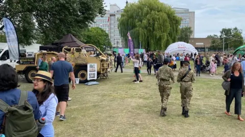 BBC Two young people, both in Army uniforms, are walking away from the camera, while in the background is an armoured vehicle, surrounded by crowds.