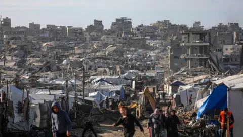EPA Internally displaced Palestinians walk among the ruins of the Al Tuffah neighborhood in eastern Gaza City, 11 February 2026, amid a ceasefire between Israel and Hamas. There are tents in front of a mountain of rubble.
