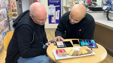 Sid Patel is sat around a small table with Rob Koch and they are looking at his phone. 