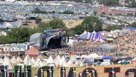 A view of the Glastonbury Festival site. There is a stage in the middle with a crowd in front of it and tents in the background. The sun is shining.